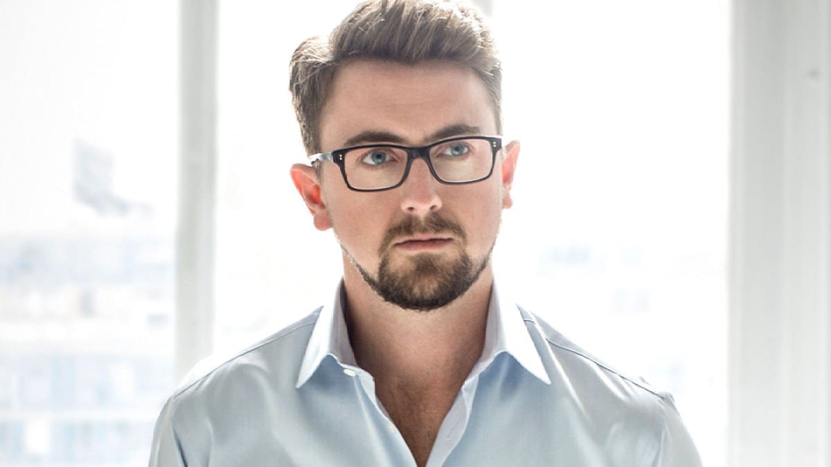 Tenor Ben Bliss, a white man wearing glasses and a light-colored shirt, stands in front of a window with natural light illuminating the scene.