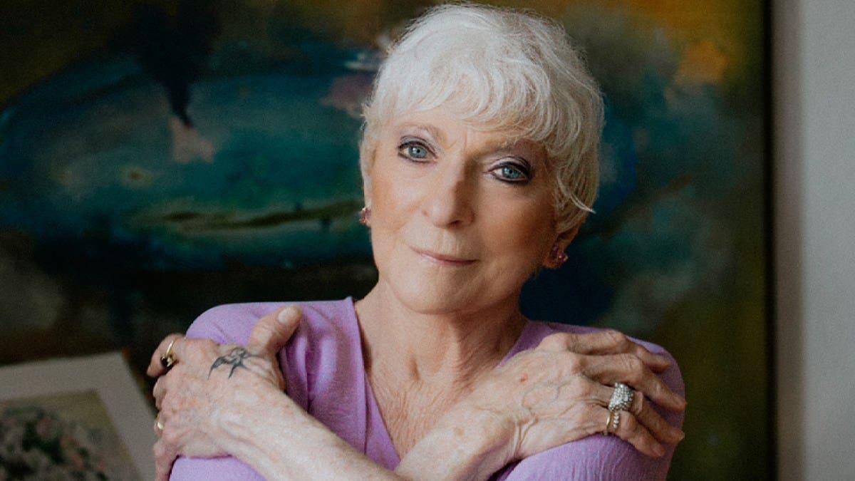 Folk singer Judy Collins, an older woman with white hair and visible hand tattoos wearing a light purple top against a colorful backdrop, looks into the camera.