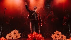 A man dressed in a mariachi outfit performs on stage, adorned with flowers and bathed in red light, celebrating the culture of Mariachi Herencia de México.