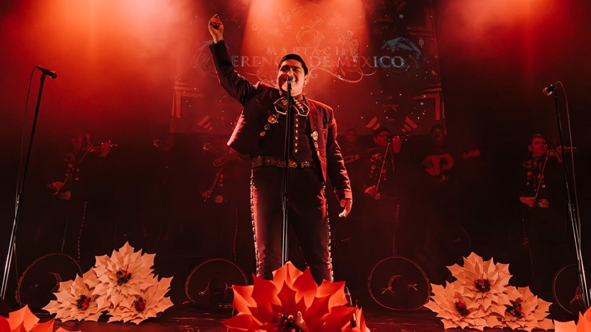 A man dressed in a mariachi outfit performs on stage, adorned with flowers and bathed in red light, celebrating the culture of Mariachi Herencia de México.