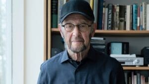 Composer Steve Reich, an older man with glasses and a baseball hat stands in front of a bookcase filled with books.
