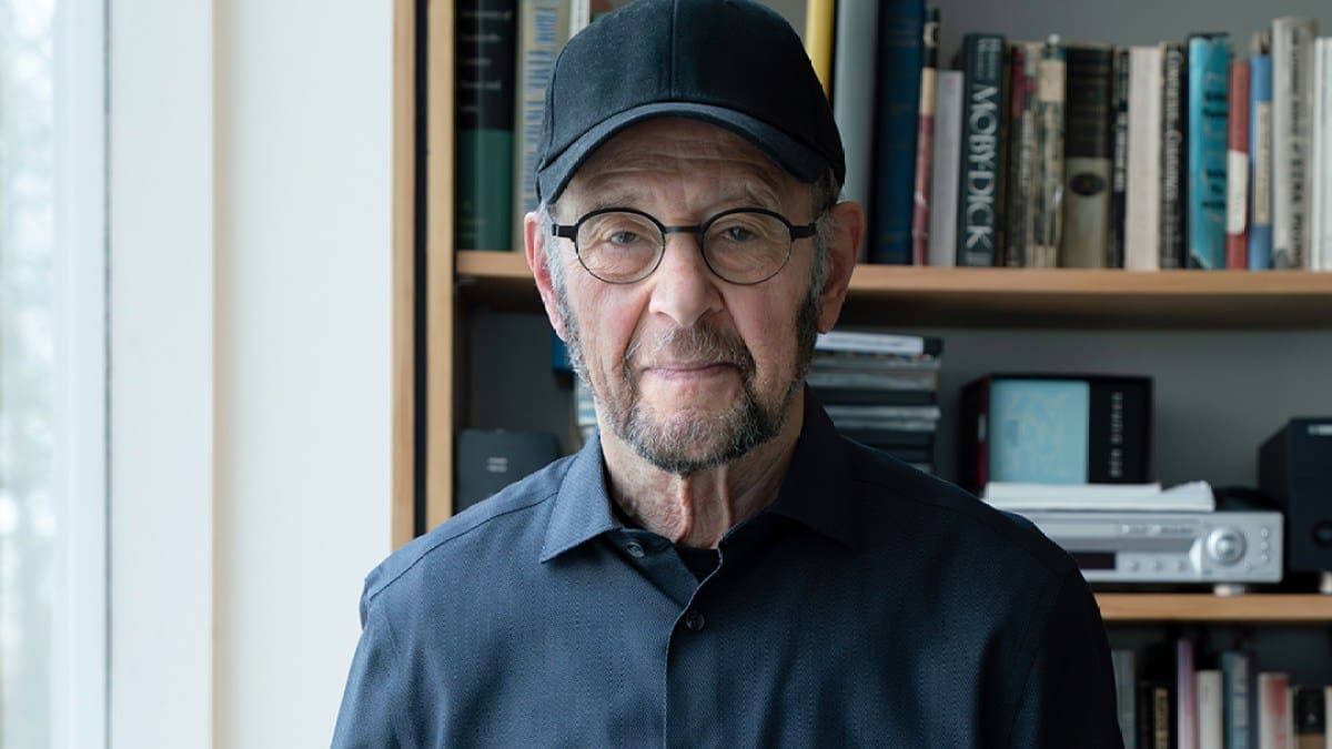 Composer Steve Reich, an older man with glasses and a baseball hat stands in front of a bookcase filled with books.
