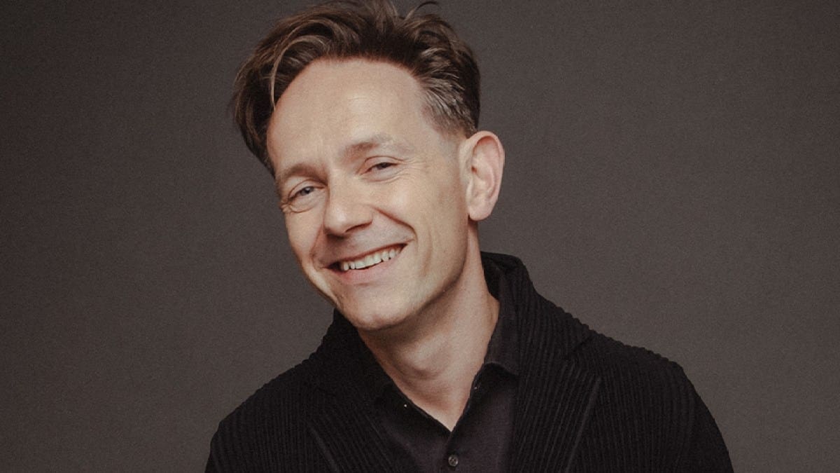 Countertenor Iestyn Davies, a smiling white man dressed in a black shirt and jacket, poses looks into the camera