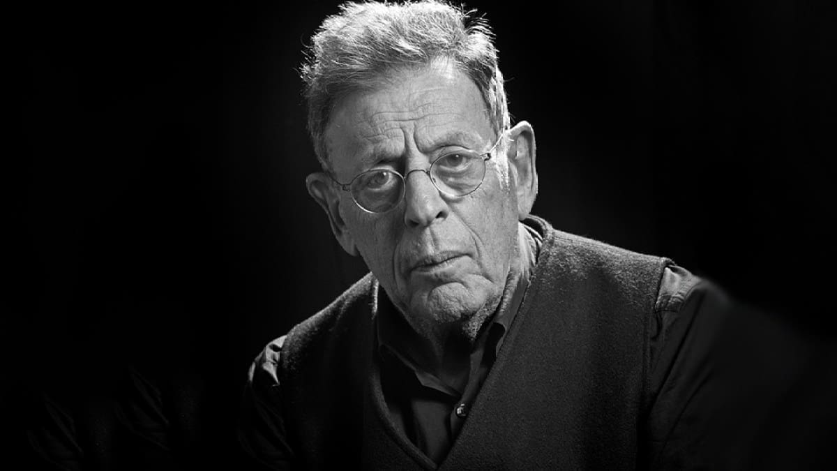 A black and white portrait of Philip Glass, an older man in a dark shirt and vest wearing glasses looking directly into the camera.