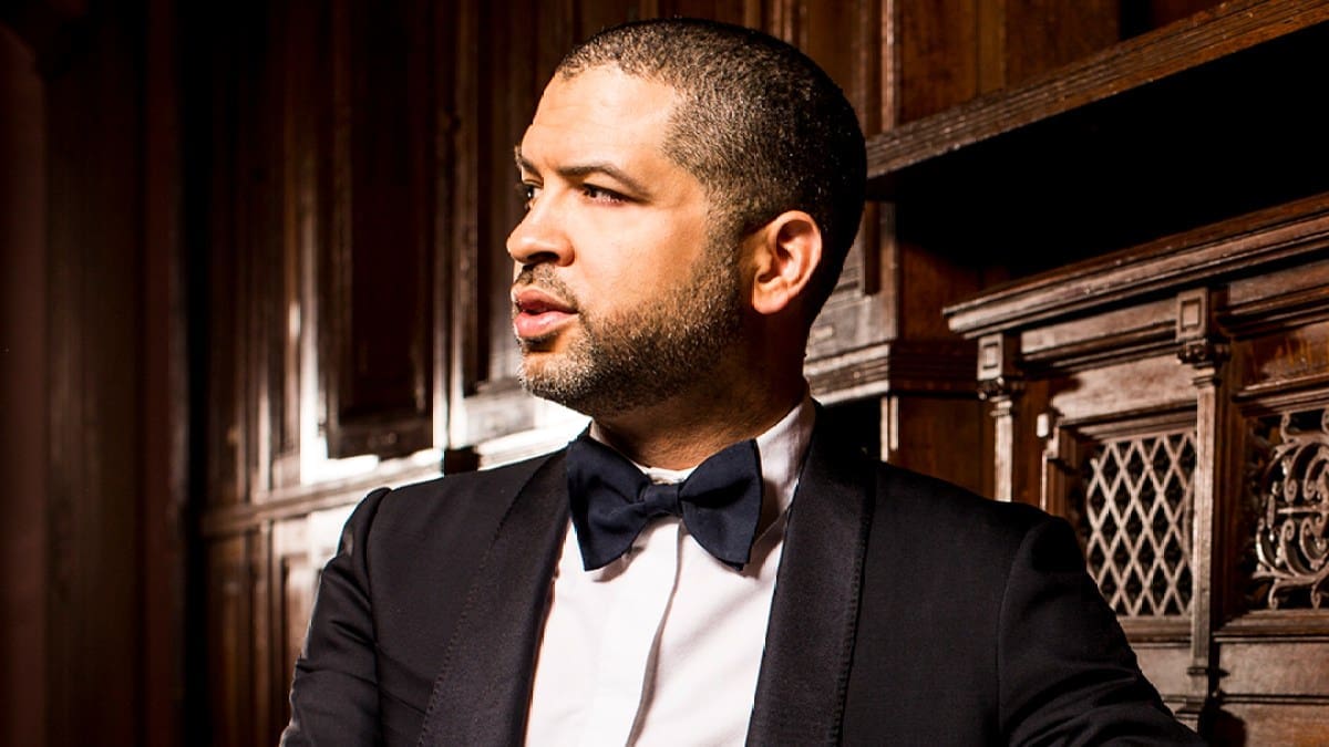 Jazz pianist Jason Moran, a black man in a black suit and bow tie, looks to the side and leans agaist an upright piano.
