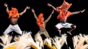 Three members -- two men and one woman -- of Cirque Kalabanté with bright orange transparent cloths over their heads and leaping into the air.