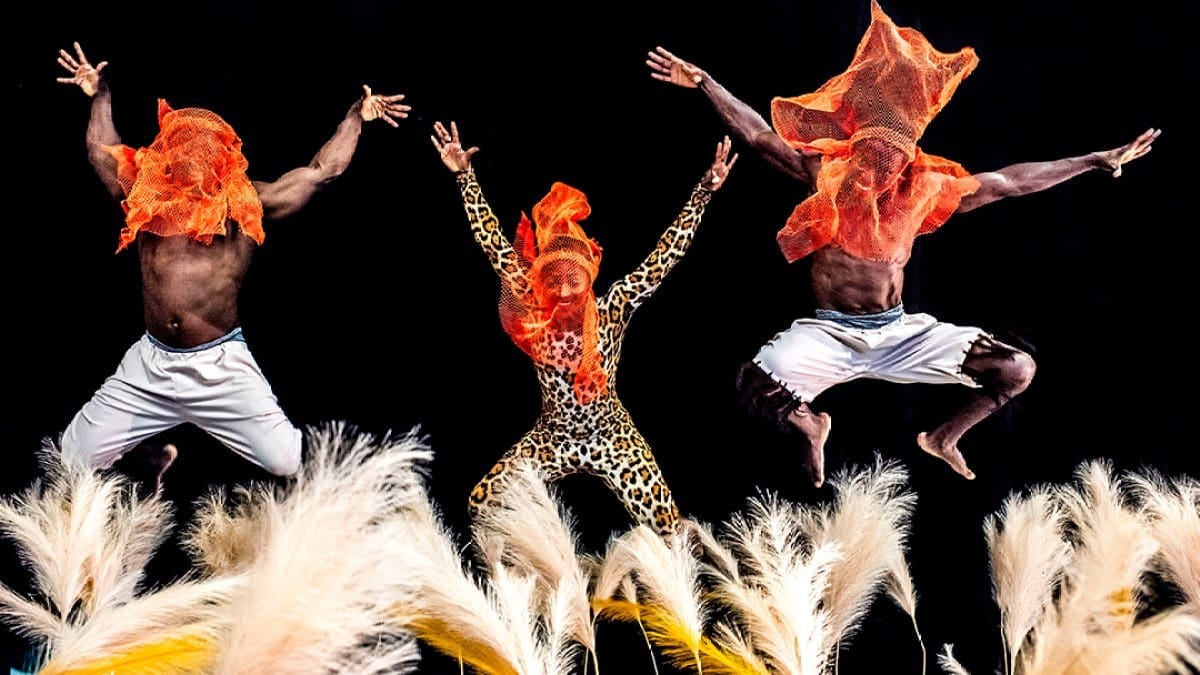 Three members -- two men and one woman -- of Cirque Kalabanté with bright orange transparent cloths over their heads and leaping into the air.