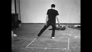 Black-and-white image of Bruce Nauman performing inside a taped square.