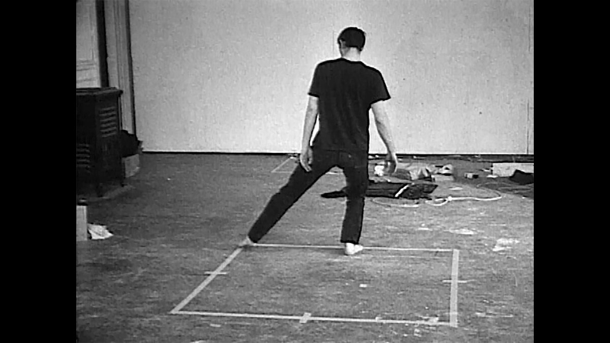 Black-and-white image of Bruce Nauman performing inside a taped square.