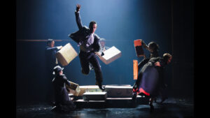 Six performers pose with luggage as a man leaps with one hand raised.