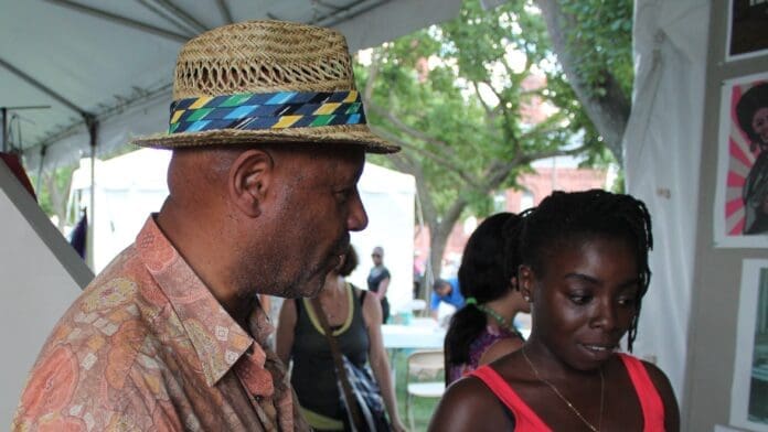 Emory Douglas, wearing a straw hat and patterned shirt, talks with a visitor beside displayed artworks.