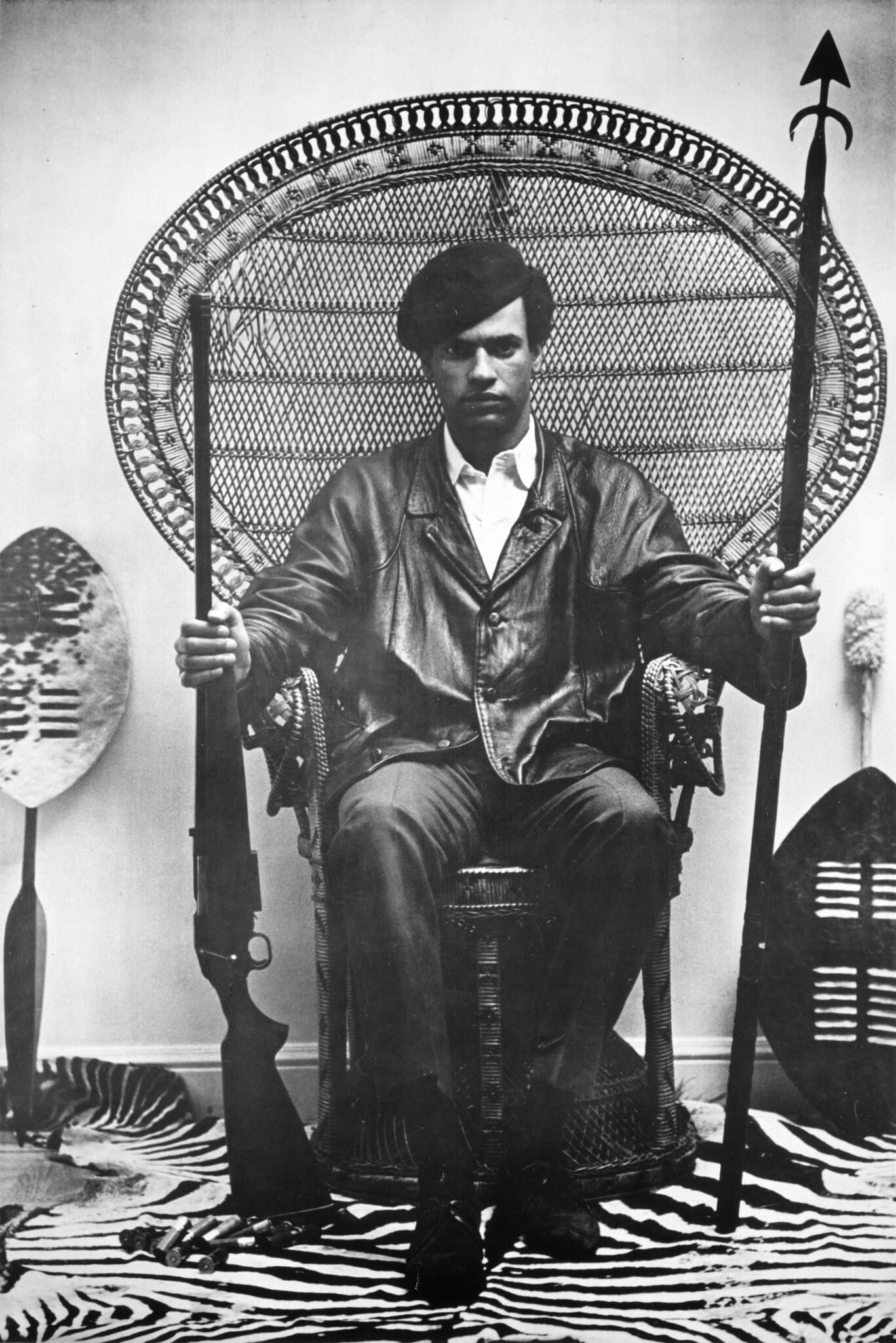 Huey P. Newton seated in a wicker chair holding a rifle and spear in Blair Stapp’s 1967 portrait